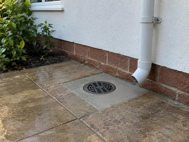 Outdoor drain grate beside brick and rendered wall with wet paving and a downpipe in Roseville